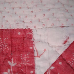 Op de afbeelding: Een doorgestikt textiel met een patchworkontwerp van afwisselende witte stoffen panelen bedrukt met kleine rode patronen en effen rode stoffen panelen. De witte secties tonen vage rode ankermotieven en delicate bloemenprints, verbonden door zichtbare witte stiksels.