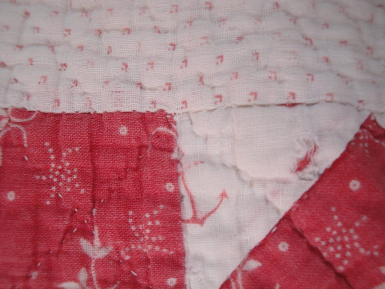 Op de afbeelding: Een doorgestikt textiel met een patchworkontwerp van afwisselende witte stoffen panelen bedrukt met kleine rode patronen en effen rode stoffen panelen. De witte secties tonen vage rode ankermotieven en delicate bloemenprints, verbonden door zichtbare witte stiksels.