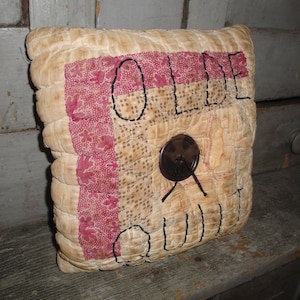 May include: A beige and red quilted pillow with the word "OLDE" embroidered in black thread. The pillow has a brown button in the center.