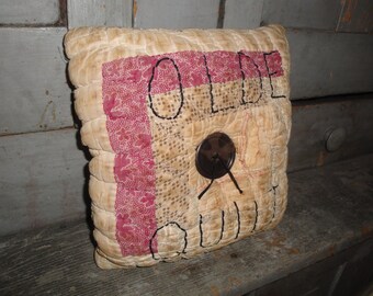 May include: A beige and red quilted pillow with the word "OLDE" embroidered in black thread. The pillow has a brown button in the center.