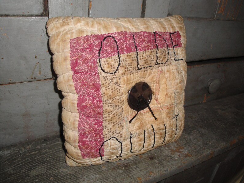 May include: A beige and red quilted pillow with the word "OLDE" embroidered in black thread. The pillow has a brown button in the center.