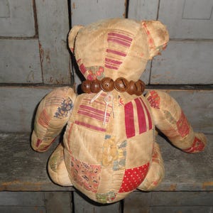 May include: A patchwork teddy bear with a brown bead necklace. The bear is sitting on a wooden surface.