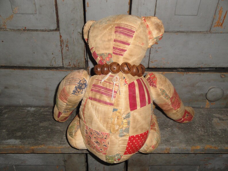 May include: A patchwork teddy bear with a brown bead necklace. The bear is sitting on a wooden surface.