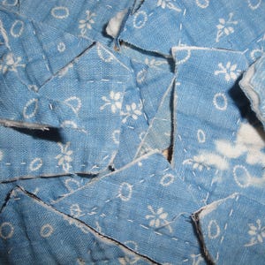 May include: A collection of light blue fabric scraps featuring a white floral and circular pattern. The pieces are quilted with visible white stitching and have raw, cut edges, revealing a white batting layer between the layers of blue fabric.