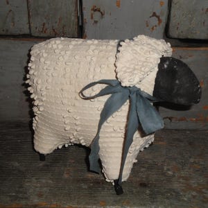 May include: A decorative sheep figurine with a textured, off-white body and a black face. It has a blue-gray ribbon tied around its neck and stands on black legs. The sheep is a rustic, country-style decorative item.