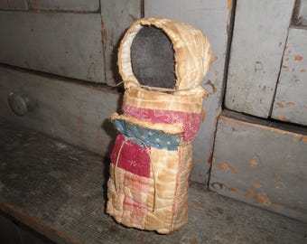May include: A handmade doll with a quilted body and head covering. The doll's body is made of patchwork fabric in shades of red, blue, and cream. The head is a dark color, and the doll has a simple, folk art style. The doll is standing on a wooden surface.