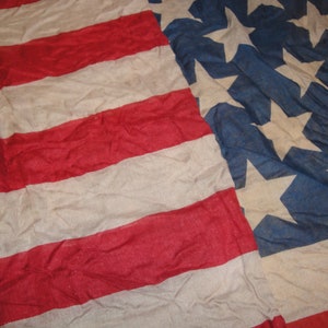 Op de afbeelding: Een close-up van een vervaagde Amerikaanse vlag met rode en witte strepen en blauwe sterren. De vlag is gemaakt van een dunne, geweven stof en heeft een versleten, vintage uitstraling.