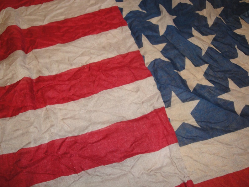 Op de afbeelding: Een close-up van een vervaagde Amerikaanse vlag met rode en witte strepen en blauwe sterren. De vlag is gemaakt van een dunne, geweven stof en heeft een versleten, vintage uitstraling.