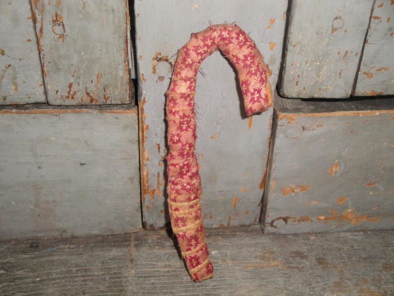 K&ouml;nnte beinhalten: Ein mit Stoff bezogener Zuckerstangen-Anh&auml;nger mit tiefrotem Hintergrund und einem sich wiederholenden goldenen Blumenmuster. Das gebogene Objekt ist mit Textilstreifen umwickelt, wobei ausgefranste Kanten an den N&auml;hten sichtbar sind.