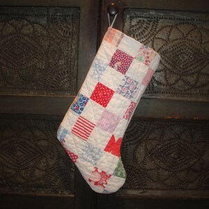 May include: A white quilted Christmas stocking with red, blue, and white patchwork squares. The stocking is hanging on a rustic wooden door.
