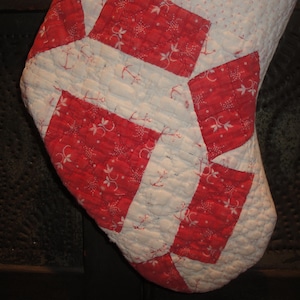 Op de afbeelding: Een gewatteerde kerstsok met een geometrisch patchworkontwerp. De stof wisselt af tussen een witte achtergrond met kleine blauwe stippen en rode rechthoekige vlakken versierd met een wit bloemen- en ankerpatroon.
