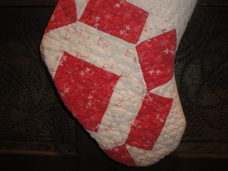 Op de afbeelding: Een gewatteerde kerstsok met een geometrisch patchworkontwerp. De stof wisselt af tussen een witte achtergrond met kleine blauwe stippen en rode rechthoekige vlakken versierd met een wit bloemen- en ankerpatroon.