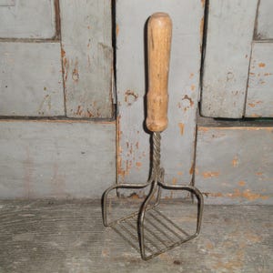 May include: Vintage potato masher with a wooden handle and a metal wire head. The handle is light brown, and the metal is a silver color. The masher is designed with a rectangular grid for mashing potatoes or other soft foods.