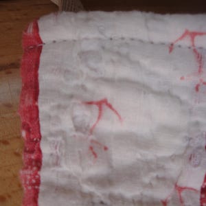 Op de afbeelding: Een zachte, meerlaagse witte katoenen stof met een gekreukte textuur, voorzien van een rode rand en verspreide rode ankermotieven. Het materiaal is langs de randen gestikt en toont een licht, ademend weefsel met zichtbare plooien.