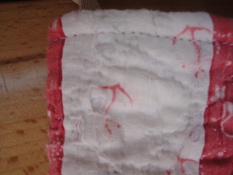 Op de afbeelding: Een zachte, meerlaagse witte katoenen stof met een gekreukte textuur, voorzien van een rode rand en verspreide rode ankermotieven. Het materiaal is langs de randen gestikt en toont een licht, ademend weefsel met zichtbare plooien.