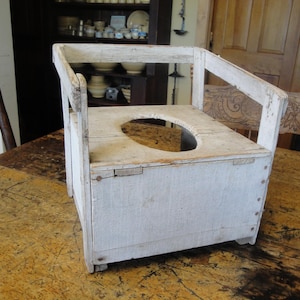 May include: A white, antique wooden commode chair with a rectangular base and a seat with a circular opening. The chair has armrests and a backrest, and it appears to be made of weathered wood. The chair is placed on a wooden table.