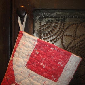 Op de afbeelding: Een gewatteerde ovenwant met een rood-wit ruitpatroon en kleine bloemmotieven. De want is geconstrueerd met een rasterachtig stikselontwerp en bevat een witte stoffen lus aan de bovenkant om hem op te hangen.