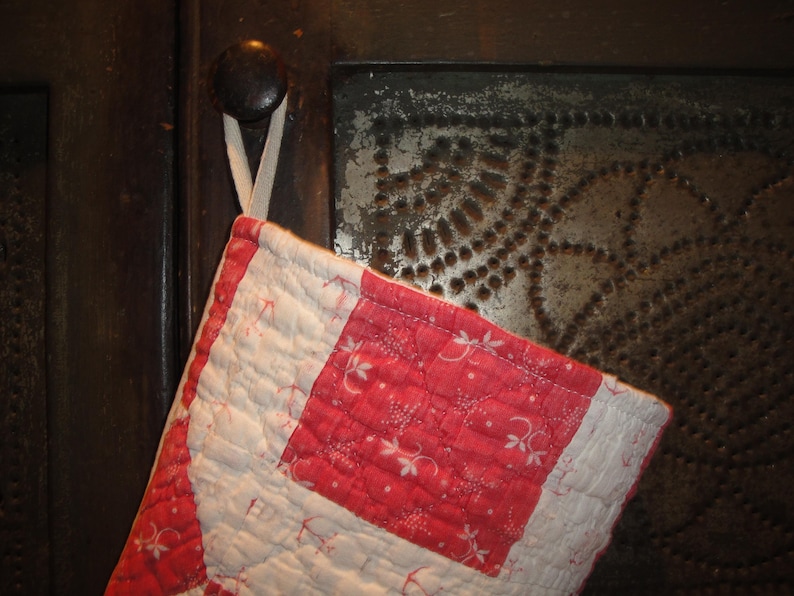 Op de afbeelding: Een gewatteerde ovenwant met een rood-wit ruitpatroon en kleine bloemmotieven. De want is geconstrueerd met een rasterachtig stikselontwerp en bevat een witte stoffen lus aan de bovenkant om hem op te hangen.