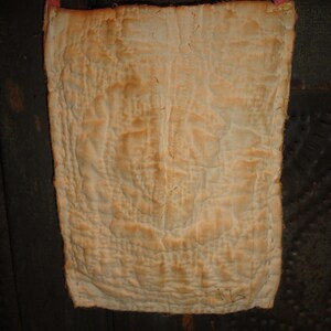 Op de afbeelding: Een rechthoekige, gewatteerde tas in een vervaagde, gebroken witte kleur, met een roze band. De stof heeft een getextureerde, gerimpelde uitstraling met donkerdere plekken. De tas lijkt handgemaakt en vintage.