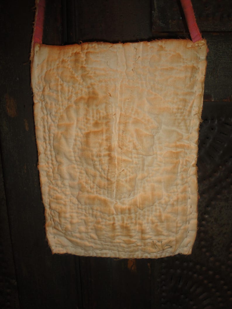 Op de afbeelding: Een rechthoekige, gewatteerde tas in een vervaagde, gebroken witte kleur, met een roze band. De stof heeft een getextureerde, gerimpelde uitstraling met donkerdere plekken. De tas lijkt handgemaakt en vintage.