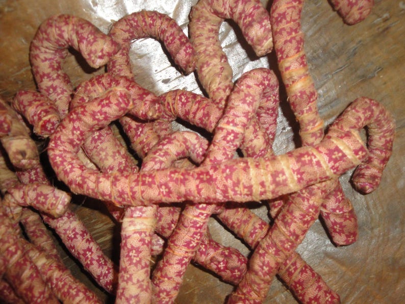 K&ouml;nnte beinhalten: Eine Ansammlung von gebogenen, stoffbezogenen St&auml;ben mit einem sich wiederholenden Muster aus kleinen roten Blumenformen auf beigem Grund. Jeder Stab ist mit einem durchgehenden Streifen aus bedrucktem Textil umwickelt, was eine texturierte, zylindrische Optik erzeugt.