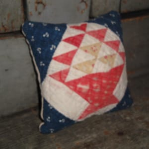 May include: Small, square pillow with a patchwork design. The pillow features a blue background with white dots and a central design of red, white, and tan geometric shapes, resembling a basket. The pillow is placed on a wooden surface.
