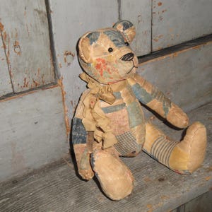 Puede incluir: Un oso de peluche confeccionado con varios parches de tela en tonos apagados de azul, beige y patrones florales rojos. El oso presenta una nariz cosida, ojos oscuros y una cinta de tela deshilachada atada alrededor del cuello, con costuras visibles.