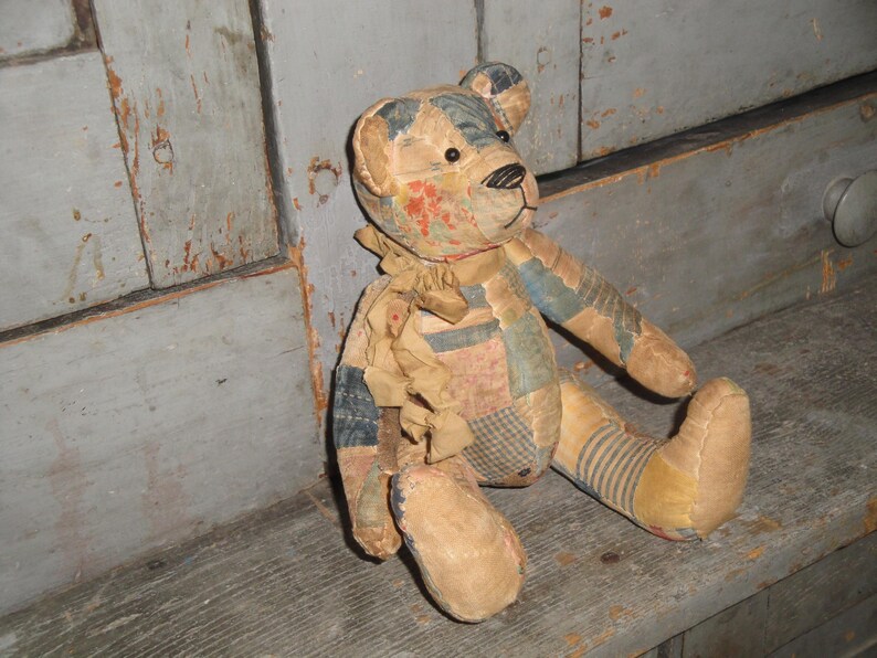 Puede incluir: Un oso de peluche confeccionado con varios parches de tela en tonos apagados de azul, beige y patrones florales rojos. El oso presenta una nariz cosida, ojos oscuros y una cinta de tela deshilachada atada alrededor del cuello, con costuras visibles.
