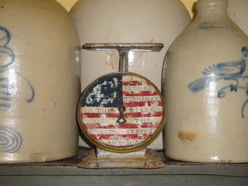 May include: A vintage scale with a red, white, and blue American flag design. The scale reads "I pledge allegiance to the flag of the United States of America".