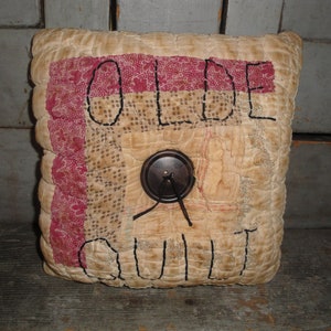 May include: A beige and red patchwork pillow with the words "Olde Quilt" embroidered on it. A brown button with a black string is in the center of the pillow.