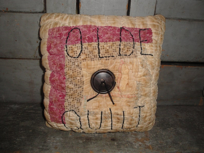 May include: A beige and red patchwork pillow with the words "Olde Quilt" embroidered on it. A brown button with a black string is in the center of the pillow.
