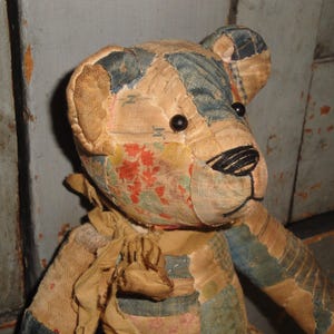 Puede incluir: Un oso de peluche de patchwork hecho de varios retazos de tela en tonos beige, azul y estampados florales. El oso presenta peque&ntilde;os ojos de cuentas negras, una nariz y boca cosidas a mano con hilo negro y una cinta de tela beige deshilachada atada al cuello.