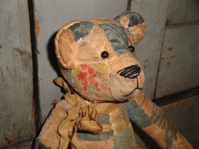 Puede incluir: Un oso de peluche de patchwork hecho de varios retazos de tela en tonos beige, azul y estampados florales. El oso presenta peque&ntilde;os ojos de cuentas negras, una nariz y boca cosidas a mano con hilo negro y una cinta de tela beige deshilachada atada al cuello.