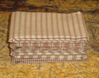 May include: A stack of folded fabric napkins in various patterns. The top napkin features vertical stripes in a muted red hue against a beige background. Below, other napkins display plaid and checkered designs in similar colors. The napkins are neatly stacked.