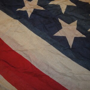 Op de afbeelding: Een close-up van een vervaagde Amerikaanse vlag met rode, witte en blauwe strepen en witte sterren op een blauw veld. De vlag is gemaakt van een dunne, geweven stof en heeft een versleten, vintage uitstraling.