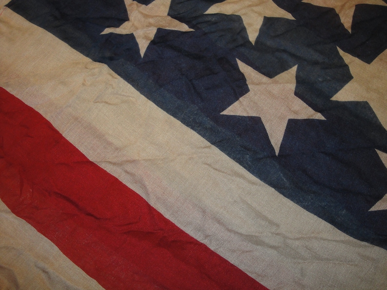 Op de afbeelding: Een close-up van een vervaagde Amerikaanse vlag met rode, witte en blauwe strepen en witte sterren op een blauw veld. De vlag is gemaakt van een dunne, geweven stof en heeft een versleten, vintage uitstraling.