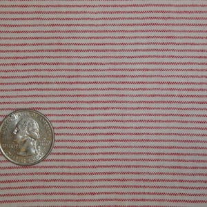 May include: Close-up of a fabric with a repeating pattern of thin, horizontal red stripes on a cream background. A U.S. quarter coin is in the lower left corner for scale. The fabric appears to be cotton or a similar woven material.