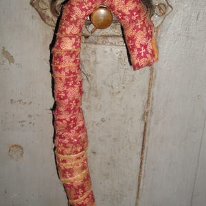 K&ouml;nnte beinhalten: Ein zuckerstangenf&ouml;rmiges Ornament, das mit rotem Stoff mit einem sich wiederholenden goldenen Blumenmuster umwickelt ist. Der Stoff ist in &uuml;berlappenden Streifen um die Form gewickelt, was eine strukturierte Oberfl&auml;che erzeugt. Das Ornament ist oben gebogen.