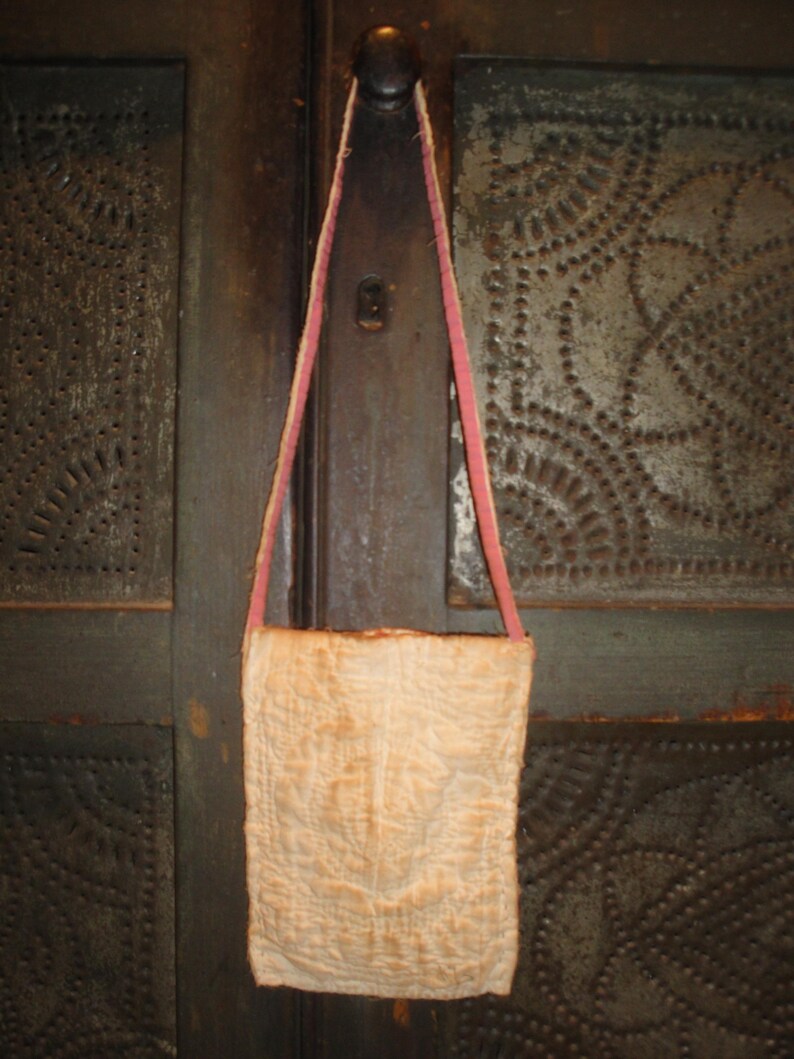 Op de afbeelding: Een cr&egrave;mekleurige, gewatteerde stoffen tas met een roze band hangt tegen een donkere, getextureerde achtergrond. De tas heeft een rechthoekige vorm en een eenvoudig ontwerp, wat wijst op een vintage of handgemaakte esthetiek. De band is dun en lijkt van een zacht materiaal te zijn.