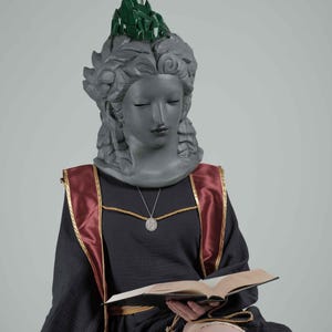 Puede incluir: Estatua de una mujer con los ojos cerrados, vestida con un vestido negro con detalles rojos y dorados, y una corona de cristal verde. Sostiene un libro abierto en su regazo, con un collar con colgante plateado.