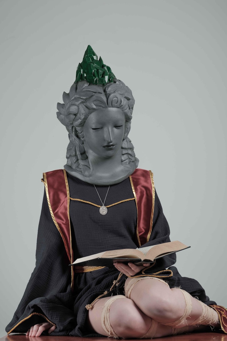 Puede incluir: Estatua de una mujer con los ojos cerrados, vestida con un vestido negro con detalles rojos y dorados, y una corona de cristal verde. Sostiene un libro abierto en su regazo, con un collar con colgante plateado.