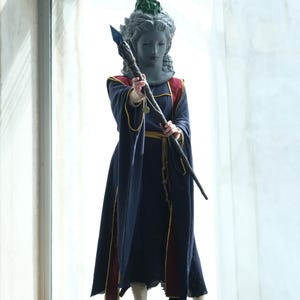 Puede incluir: Una persona con una cabeza de estatua gris con una corona verde y una t&uacute;nica azul marino con detalles rojos y dorados. La persona sostiene un bast&oacute;n con punta azul.
