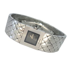 May include: A silver-toned wristwatch featuring a square black dial with two hands and the text CHANEL and SWISS. The band is constructed from interconnected metallic segments forming a quilted pattern that wraps around the wrist.