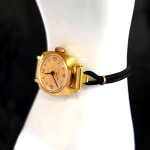 May include: A gold-tone analogue wristwatch with a round face, featuring a light-coloured dial with black numerals at the 3, 6, 9, and 12 positions. The watch has a thin black cord strap secured with a small gold-tone metal ring connector.