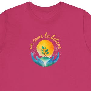 May include: A magenta t-shirt featuring a watercolor design. The design includes a yellow sun, a green tree, and blue hands cradling the tree. The text "we came to believe" is written in yellow script above the sun.