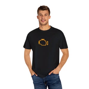 Może przedstawiać: Czarny t-shirt z żółtym symbolem silnika. Grafika jest wyśrodkowana na klatce piersiowej. Koszulka ma okrągły dekolt i jest wykonana z miękkiego materiału. Model ma na sobie niebieskie dżinsy.