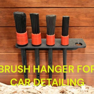 May include: A black plastic brush holder with five black brushes with red handles. The holder is mounted on a wooden wall. The text "BRUSH HANGER FOR CAR DETAILING" is printed below the holder.