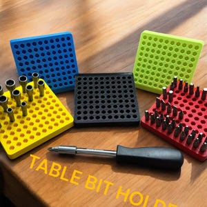May include: A set of four table bit holders in different colors: blue, yellow, black, and red. Each holder has a grid of hexagonal holes to store various bits. The holders are arranged on a wooden surface with a screwdriver in the foreground. The text "TABLE BIT HOLDER" is written in yellow letters on the wooden surface.