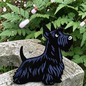 May include: An embroidered patch shaped like a Scottish Terrier dog. The dog is black with dark blue highlights in the fur, pointed ears, a short upright tail, and a white eye and nose detail. The embroidery features a textured, layered stitch.