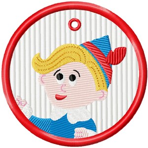May include: Embroidered design of a cartoon character with blonde hair, a blue hat with red accents, and a blue outfit. The design is framed by a red circle with a small loop at the top, set against a white striped background.